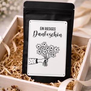 Könnte beinhalten: Ein schwarzer Standbeutel mit einem weißen rechteckigen Etikett, das den Text "EIN RIESIGES Dankeschön" und eine schwarze Strichzeichnung einer Hand mit einem Blumenstrauß zeigt, platziert in einer Holzkiste mit hellen Holzspänen.
