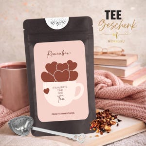Könnte beinhalten: Ein schwarzer Teebeutel mit einem hellrosa Etikett, das eine weiße Teetasse mit braunen Herzen zeigt. Der Text lautet Remember: IT'S ALWAYS TIME FOR Tea und FRÜCHTETEEMISCHUNG. Ein silbernes herzförmiges Teesieb liegt vor dem Beutel.
