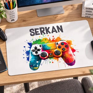 Könnte beinhalten: Eine rechteckige Schreibtischunterlage mit weißem Hintergrund und dem in Schwarz gedruckten Namen SERKAN über einer farbenfrohen Grafik eines Videospiel-Controllers im Farbspritzer-Stil. Das Controller-Design enthält lebendige Regenbogenfarben.