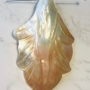 Puede incluir: Un gran colgante de concha nacarada blanco y melocotón con un diseño tallado. La concha tiene forma de hoja y tiene un agujero en la parte superior para colgar.