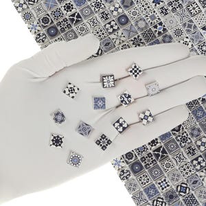 May include: A collection of small, square decorative tiles with intricate blue and white patterns, held in a white gloved hand. The tiles feature various geometric and floral designs, with a matching patterned background.