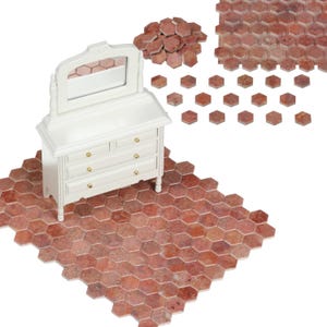 Puede incluir: Un tocador blanco en miniatura con espejo, sobre un suelo de baldosas hexagonales rojas. Hay baldosas rojas adicionales esparcidas alrededor del mueble. El tocador tiene tres cajones con pomos dorados.