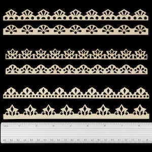 May include: Six light-coloured wooden laser-cut border strips with various geometric and fan-shaped patterns, arranged horizontally above a clear plastic ruler that measures up to 20 cm in length, showing the scale of the wooden pieces.