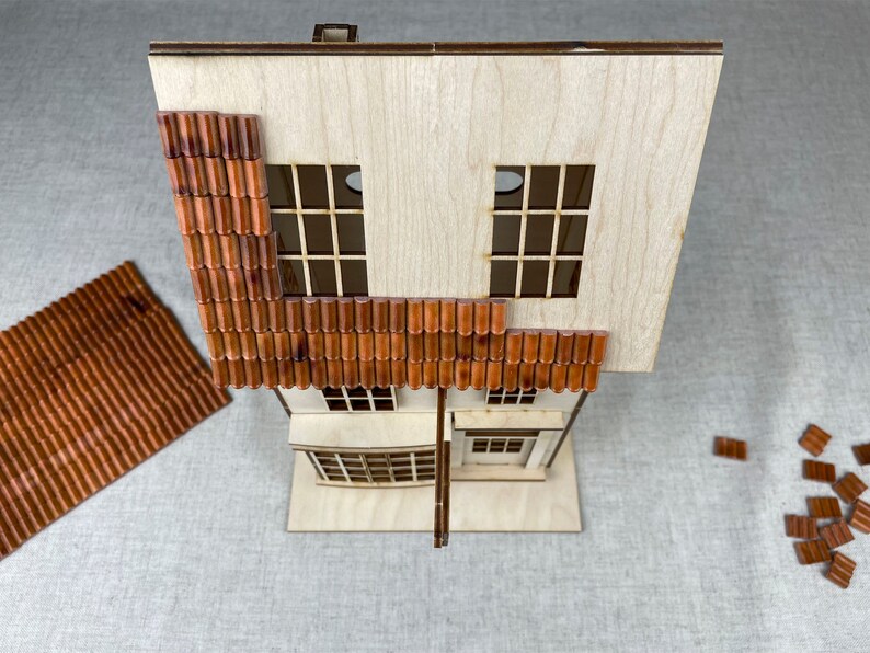 May include: A wooden model house with brown tile roofing. The house has two windows and a front door. The roof is partially assembled with some tiles missing.