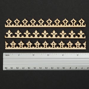 May include: Three light-colored wooden trim strips with decorative cutouts, each measuring approximately 5.5 inches in length. The strips feature repeating pointed architectural patterns and are positioned above a metal ruler showing imperial units.