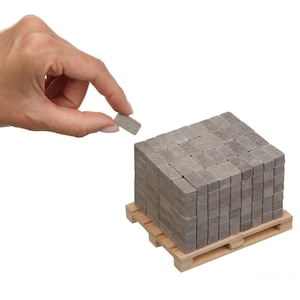 Könnte beinhalten: Eine Hand hält einen einzelnen grauen Betonpflasterstein, während eine Palette mit grauen Betonpflastersteinen auf einer Holzpalette steht.