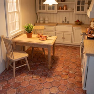 May include: A miniature dollhouse kitchen scene featuring a light wood table and chair, a small potted green plant, and two copper-colored pans. The room has terracotta-colored hexagonal floor tiles, white cabinetry, a sink, and a window.