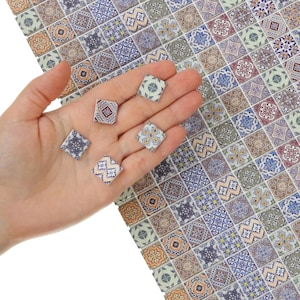 以下が含まれることがあります： 手のひらに、カラフルで複雑な模様の小さな正方形の装飾タイルがいくつかあります。タイルは、青、オレンジ、茶色のさまざまなデザインが特徴です。背景は同様のタイルのシートで、モザイク効果を生み出しています。