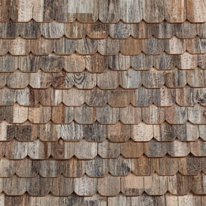 May include: Close-up of a wooden shingle roof. The shingles are a mix of brown, gray, and tan, creating a textured pattern. The individual shingles have a rounded bottom edge and are arranged in overlapping rows.