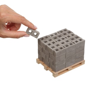 Könnte beinhalten: Eine Palette mit Miniatur-Betonsteinen, jeder mit einem quadratischen Loch in der Mitte. Die Steine sind ordentlich gestapelt und haben eine hellgraue Farbe. Ein einzelner Stein wird in der Abbildung von einer Hand gehalten.