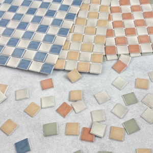 May include: Assortment of small ceramic tiles in various colors, including blue, beige, brown, and green. The tiles are arranged in a checkered pattern and are scattered on a light gray surface.