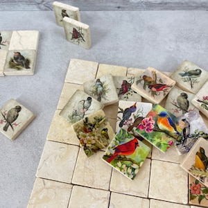 May include: A set of square ceramic tiles with images of birds on them. The tiles are arranged in a grid pattern on a light beige surface. The birds are depicted in various colors and poses, including a red cardinal, a bluebird, and a sparrow.
