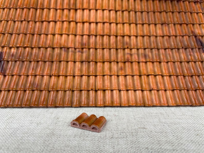 May include: A close-up of a miniature terracotta roof tile, with a textured surface and a reddish-brown color. The tile is sitting on a light-colored fabric surface.