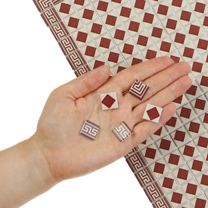May include: A hand holding several small, square tiles with a red diamond design. The tiles are surrounded by a larger surface of similar tiles, creating a decorative pattern. The tiles have a cream background with a red diamond in the center.