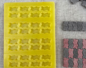 May include: A yellow silicone mould with 20 rectangular cavities. The mould is designed to create small, wavy, rectangular shapes.  There are two sets of grey and red interlocking paving stones.