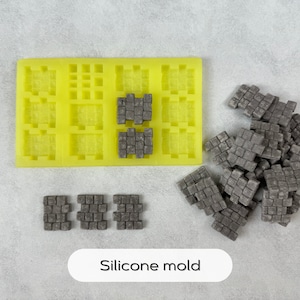 Op de afbeelding: Een gele siliconen mal met 12 individuele mallen in de vorm van kleine stenen blokken. Er zijn verschillende grijze stenen blokken die uit de mal zijn verwijderd en verspreid liggen op een wit oppervlak. De tekst "Silikon mal" is gedrukt op een witte ovale sticker.