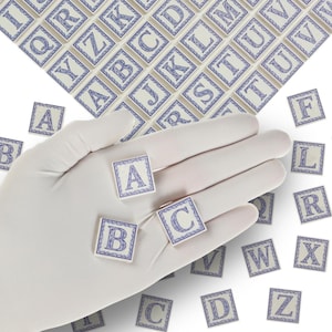 Può includere: Piastrelle quadrate con lettere dell'alfabeto in un design blu e beige. Le lettere A, B e C sono visibili sulle piastrelle tenute in una mano guantata. Altre piastrelle con lettere sono sparse.