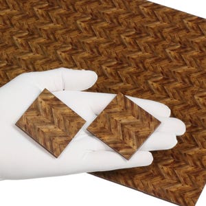 May include: Two square wooden tiles with a herringbone pattern are held in a gloved hand, with a larger sheet of matching material in the background. The tiles and sheet have a warm brown color.