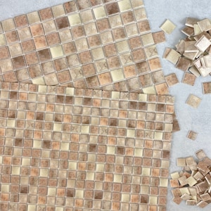May include: Overhead view of mosaic tiles in shades of beige, tan, and brown. Two sheets of tiles are intact, while a pile of individual tiles sits nearby. The tiles are square and arranged in a grid pattern, suitable for home improvement projects.