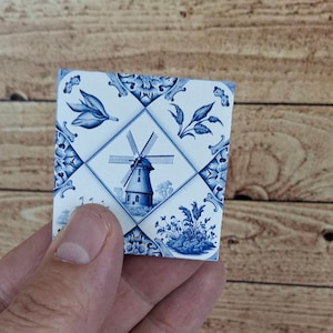 Puede incluir: Una baldosa de cerámica cuadrada con un diseño de molino de viento azul y blanco. La baldosa presenta una imagen central de molino de viento rodeada de patrones florales y geométricos. La combinación de colores es principalmente azul sobre fondo blanco.