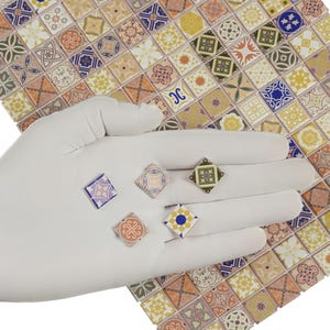 May include: A hand wearing a white glove holds five small, square decorative tiles. The tiles feature intricate patterns in various colors, including blue, orange, green, and yellow. The background shows a larger sheet of similar patterned tiles.