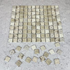 May include: A square mosaic tile sheet featuring a grid of small, square tiles in shades of beige, cream, and light brown. Some tiles have intricate, faded floral and geometric patterns, while others have a solid, weathered, or distressed finish.