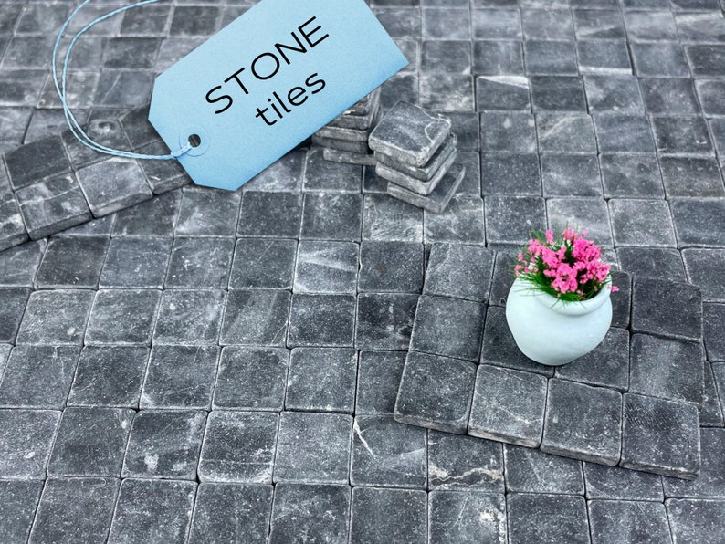 Może przedstawiać: Zbliżenie na szare kwadratowe płytki kamienne ułożone w siatkę. Niebieska etykieta z tekstem "STONE tiles" znajduje się w lewym g&oacute;rnym rogu. Mały biały pojemnik z r&oacute;żowymi kwiatami stoi na płytkach.