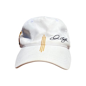 May include: White baseball cap with a curved brim. The cap features three vertical gold stripes and a signature in black on the side. The cap is made of a soft material.