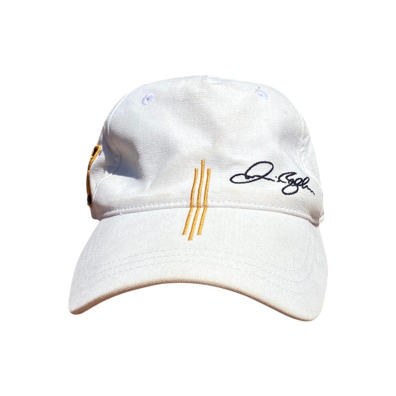 May include: White baseball cap with a curved brim. The cap features three vertical gold stripes and a signature in black on the side. The cap is made of a soft material.