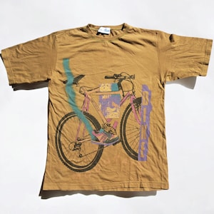 May include: A brown t-shirt with a graphic of a bicycle with a pink, blue, and purple design. The word "BIKE" is printed vertically on the right side of the shirt.