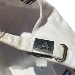 May include: White baseball cap with a metal clasp featuring the Adidas logo. The cap's adjustable strap is visible, along with the Adidas logo on the inside. The cap is made of a textured fabric.