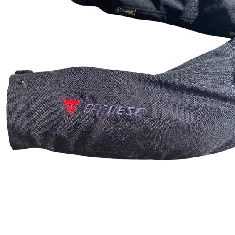 May include: Close-up of a black textile garment with the word "DAINESE" embroidered in white. A red triangle logo is above the brand name. The garment has a button and a small tag with text.