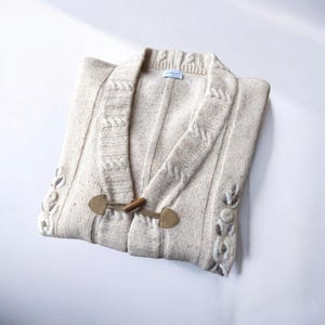 Puede incluir: Un cárdigan de punto color crema con un patrón de punto trenzado y detalles florales bordados. El cárdigan tiene cuello en V y cierre de botón.