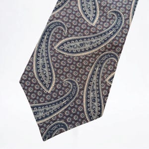 Puede incluir: Una corbata con estampado de paisley en un esquema de colores marrón, azul y blanco. La corbata tiene un extremo puntiagudo y un patrón repetitivo de formas paisley.