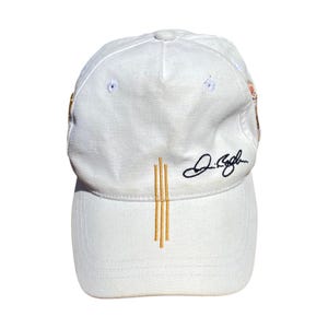 May include: A white baseball cap with a curved brim. The cap features three vertical gold lines embroidered on the front and a signature in black script. The cap is made of a soft, textured fabric.