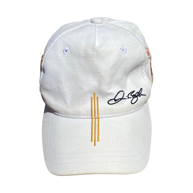 May include: A white baseball cap with a curved brim. The cap features three vertical gold lines embroidered on the front and a signature in black script. The cap is made of a soft, textured fabric.