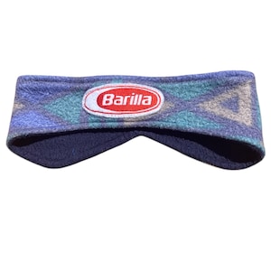 Può includere: Una fascia in pile con un motivo geometrico nei toni del blu, verde acqua e grigio. Al centro presenta una toppa ovale bianca ricamata con un bordo rosso contenente la scritta rossa Barilla. L'interno è foderato in pile blu scuro.