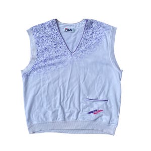 May include: White sleeveless sweater vest with a purple and pink FILA logo on the front. The vest has a purple trim around the neck and a purple pocket on the front. The vest has a faded purple and white pattern on the top half.