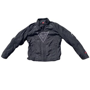 Op de afbeelding: Zwarte motorjas met een wit logo op de borst. De jas heeft een hoge kraag, ritszakken en witte accenten op de mouwen. Rode accenten zijn zichtbaar op de mouwen. De jas ligt op een witte achtergrond.