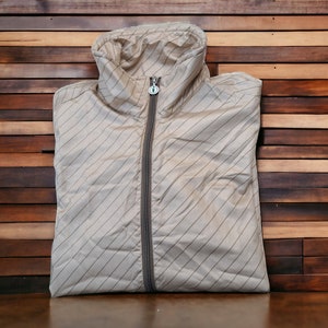 Puede incluir: Chaqueta beige con capucha y cremallera completa. La chaqueta tiene finas rayas diagonales y un pequeño logotipo en el tirador de la cremallera. La chaqueta está doblada y sobre una superficie de madera.