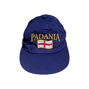 Op de afbeelding: Een marineblauwe baseballcap met het woord "PADANIA" geborduurd in gouddraad boven een vlagembleem. De vlag heeft een rood kruis op een witte achtergrond, omlijnd in goud. De cap heeft een gebogen klep.