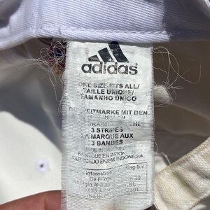 May include: Close-up of a white Adidas hat tag. The tag features the Adidas logo and text in multiple languages, including "ONE SIZE FITS ALL" and "3 STRIPES". The tag is slightly worn, with some loose threads.
