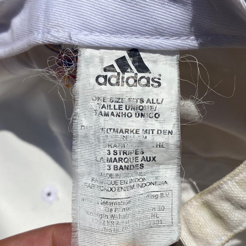 May include: Close-up of a white Adidas hat tag. The tag features the Adidas logo and text in multiple languages, including "ONE SIZE FITS ALL" and "3 STRIPES". The tag is slightly worn, with some loose threads.