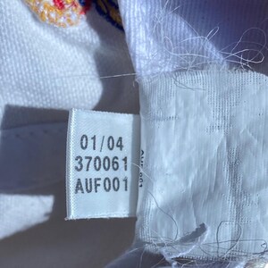 May include: Close-up of a white fabric tag with the numbers "01/04" and "370061" printed in black. Below these numbers is the text "AUF001". The tag is attached to white fabric.