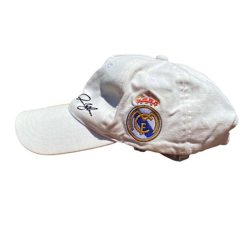 May include: White baseball cap featuring the Real Madrid logo embroidered on the side. The logo includes a crown, a circular emblem with a gold and blue design, and a signature on the side.