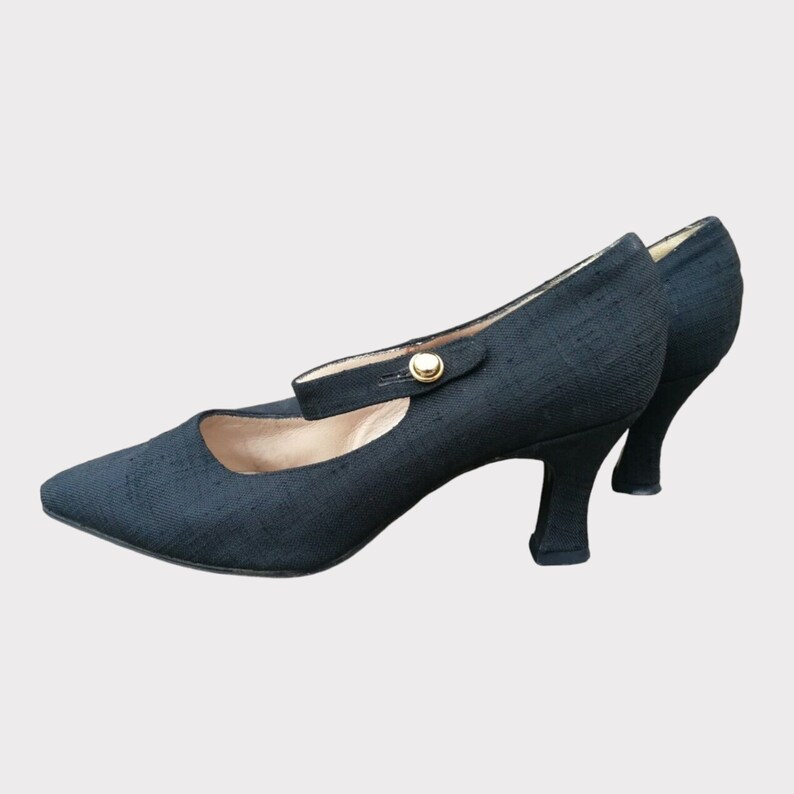 May include: Black Mary Jane heels with a pointed toe and a small heel. The shoes have a strap that fastens with a gold-colored button. The heels are approximately 2 inches (5 cm) high.