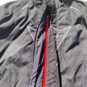 May include: Black motorcycle jacket with a red zipper and white logo accents. The jacket has a high collar and a zippered front closure. The interior lining is black. The jacket is made of a durable, weather-resistant material.