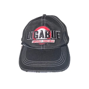 Può includere: Un cappellino da baseball nero con cuciture bianche a contrasto. La parte anteriore presenta una toppa ricamata con il testo LIGABUE in lettere nere su sfondo circolare rosso e bianco, e un'etichetta rettangolare CAMPOVOLO REGGIO EMILIA.