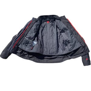 May include: Black motorcycle jacket with red accents. The jacket features a quilted black lining, multiple pockets, and a red logo on the sleeve. The jacket is open, revealing the interior details.