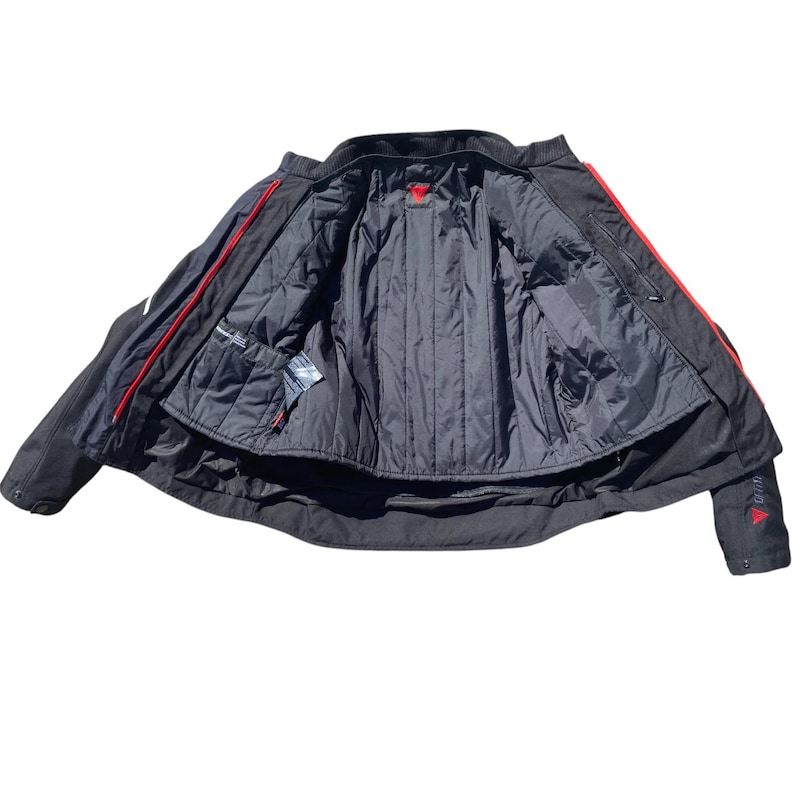 May include: Black motorcycle jacket with red accents. The jacket features a quilted black lining, multiple pockets, and a red logo on the sleeve. The jacket is open, revealing the interior details.